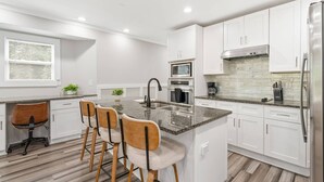 A modern kitchen with white cabinets, a granite countertop, a central island with bar stools, a stainless steel refrigerator, and a window with blinds. - Stylish 4BR Home Near ATL Airport & Beltline (Atlanta)
