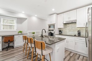 Eine moderne Küche mit weißen Schränken, einer Arbeitsplatte aus Granit, einer zentralen Kücheninsel mit Barhocker, einem Edelstahlkühlschrank und einem Fenster mit Jalousien.
