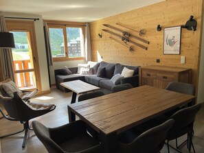 Ein gemütliches Zimmer mit einem Holztisch, einer Couch und einem Sessel. An der Wand sind Skier befestigt und ein Poster hängt dort.