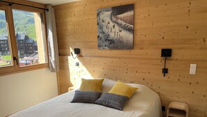 Una habitación con paneles de madera, una cama, una mesita de noche, una lámpara de pared y una ventana con vistas a edificios.
