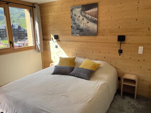 Ein Zimmer mit Holzvertäfelung, einem Bett, einem kleinen Nachttisch, einer Wandlampe und einem Fenster mit Blick auf Gebäude.
