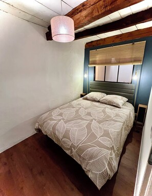 A bedroom with a bed, a headboard, a window with blinds, a nightstand, and a wall-mounted lamp. - Cozy apartment in the heart of Veynes with a balcony and a view of the mountains 🏔️ (Veynes)