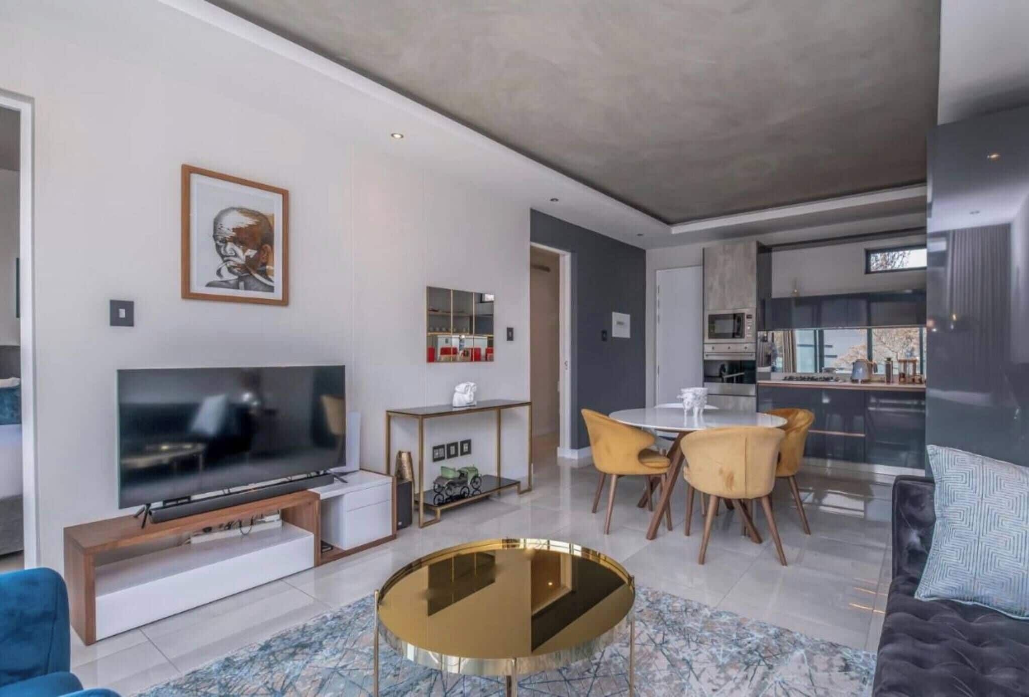 A modern living room with a flat-screen TV, a dining area with yellow chairs, and a kitchen in the background.