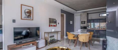A modern living room with a flat-screen TV, a dining area with yellow chairs, and a kitchen in the background.