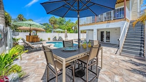 A poolside area with a table and chairs, a large umbrella, and a staircase leading to an upper level.