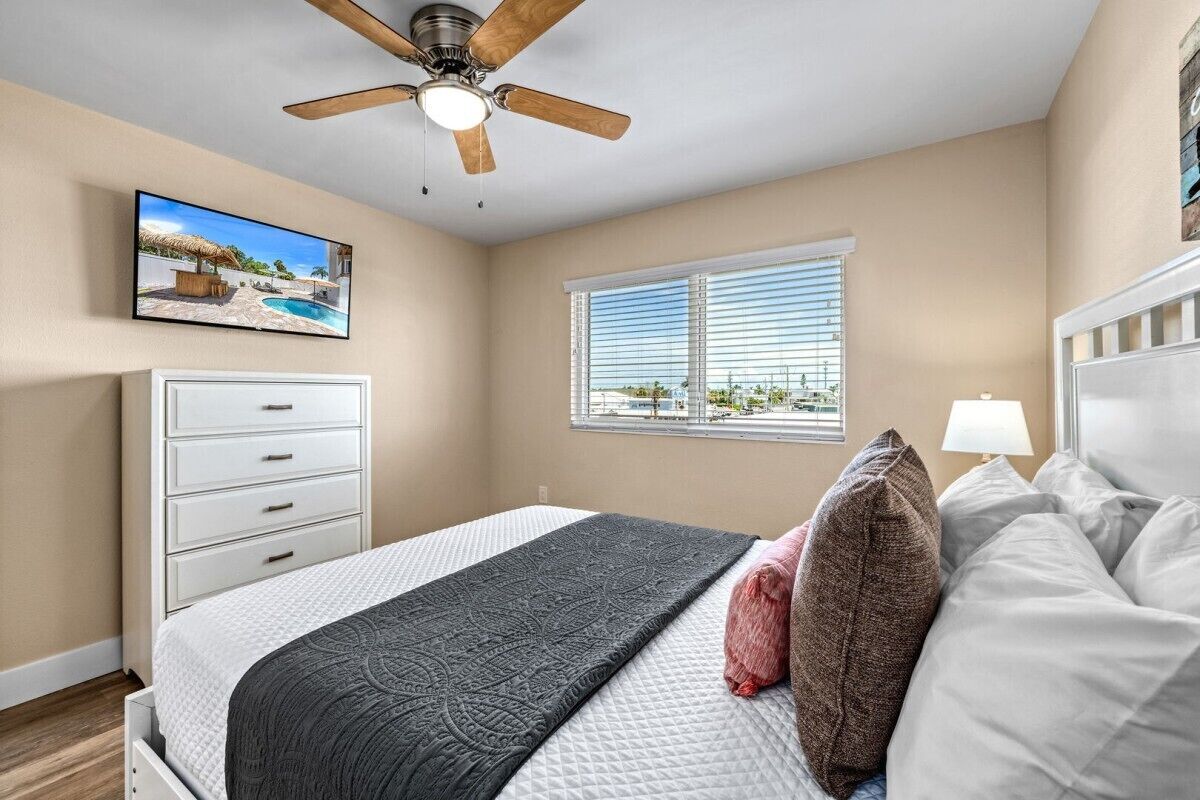 A bedroom with a bed, a ceiling fan, a window with blinds, a TV on the wall, and a view of the outdoors.