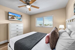 A bedroom with a bed, a ceiling fan, a window with blinds, a TV on the wall, and a view of the outdoors.