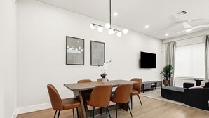 A modern dining area with a wooden table and leather chairs, a flat-screen TV on the wall, and a large window with blinds. - New Build Near Downtown + Balcony + Backyard (Houston)