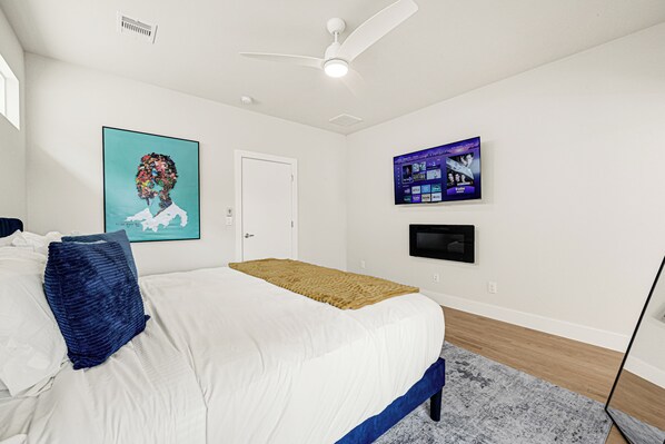 Un dormitorio con una cama, un ventilador de techo, un televisor montado y obras de arte en las paredes.