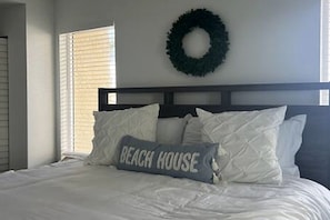 Quarto com cama, travesseiro com a inscrição 'BEACH HOUSE', um arranjo de flores na parede e janela com persianas.