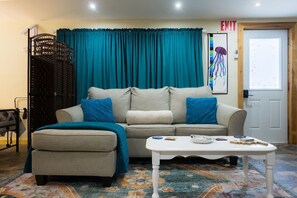 A room with a sofa, ottoman, and a coffee table. There is a door with an exit sign and a painting of a jellyfish on the wall.