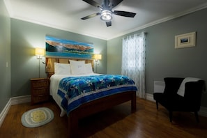 A bedroom with a bed, a nightstand, a chair, a painting on the wall, and a ceiling fan. - Fisherman's Place - Anchor Down (Rocky Harbour)