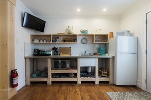 A compact kitchen with a microwave, oven, and refrigerator. There is a wooden shelf unit with various kitchen items and a clock. A fire extinguisher is mounted on the wall. - Killick Place - Anchor Down (Rocky Harbour)