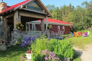 A red building with a stone facade, a porch with outdoor seating, and a garden with various plants. - Killick Place - Anchor Down (Rocky Harbour)
