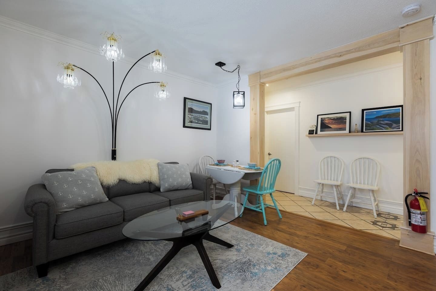 A living room with a gray sofa, a glass coffee table, and a teal chair. There are two framed pictures on the wall and a fire extinguisher near the door.