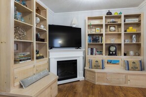 Ein Wohnzimmer mit Kamin, Holzregalen mit Büchern und Dekoration, einem Flachbildfernseher und einem kleinen Tisch mit Kissen.