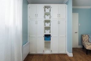 Una habitación con un armario blanco, un cesto azul para guardar cosas y una silla estampada.