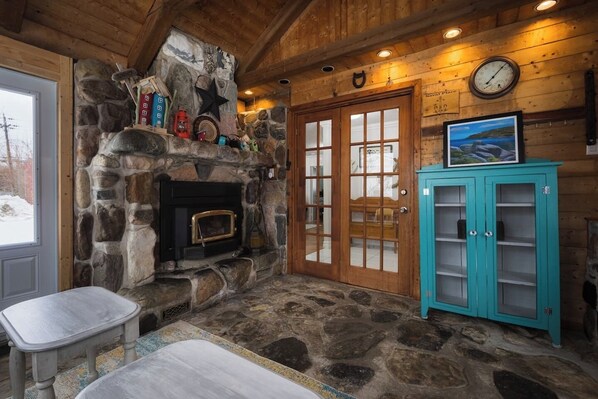 A stone fireplace with a clock and a framed picture above it, a teal cabinet with glass doors, and a wooden ceiling.