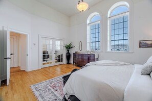 A bedroom with a large bed, a dresser, a vase with plants, and a view through the windows.