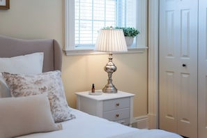 A bedroom with a bed, a bedside table with a lamp, a window with blinds, and a door.