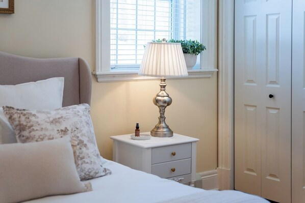 A bedroom with a bed, a bedside table with a lamp, a window with blinds, and a door.
