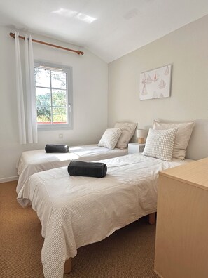 A bedroom with two beds, a window with curtains, a bedside table, and a wall-mounted artwork.