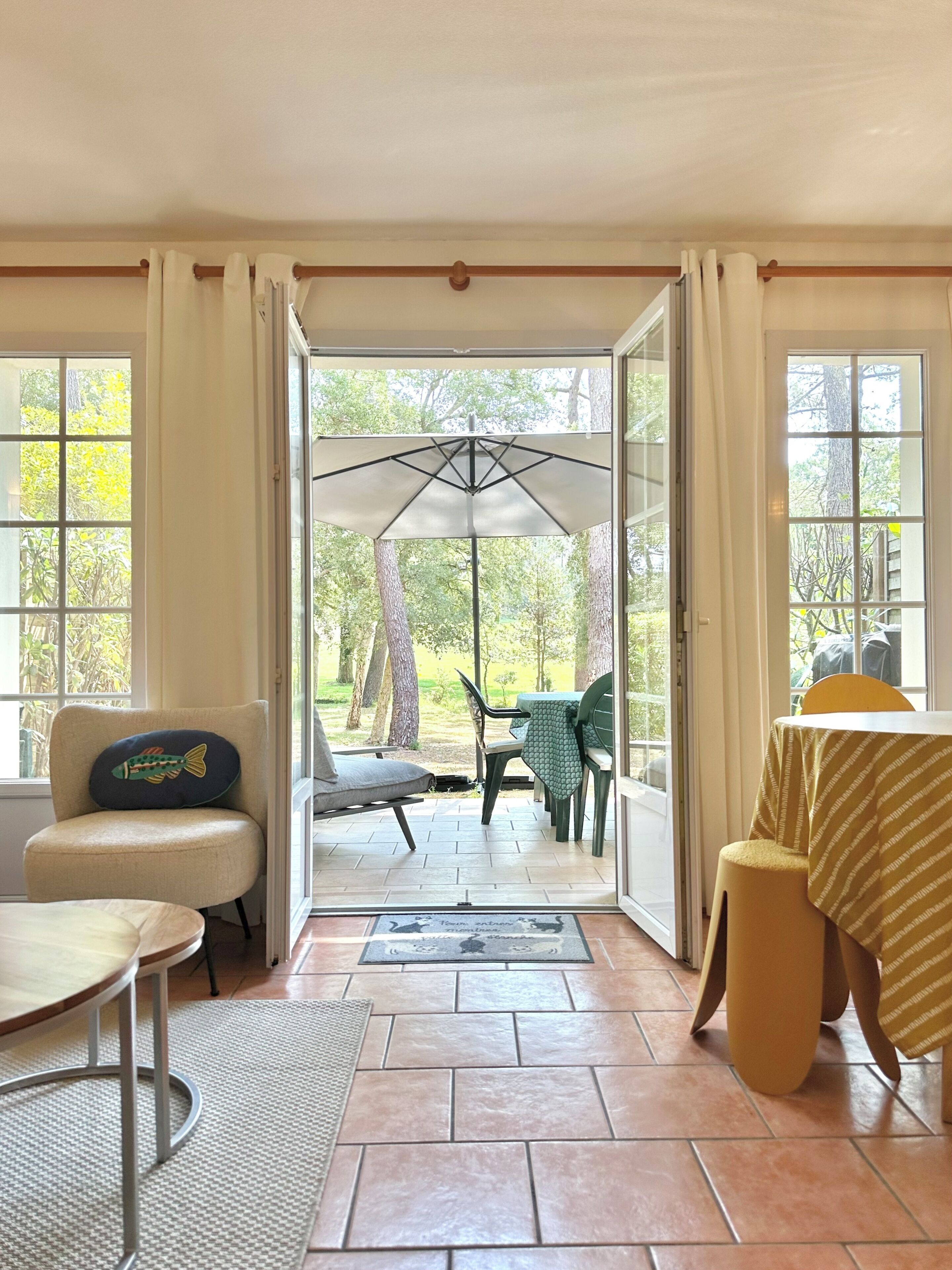 A room with a view of a patio area, a chair with a fish-shaped pillow, a table with a striped tablecloth, and a door leading outside.