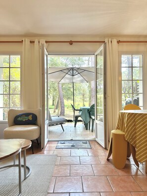 A room with a view of a patio area, a chair with a fish-shaped pillow, a table with a striped tablecloth, and a door leading outside.