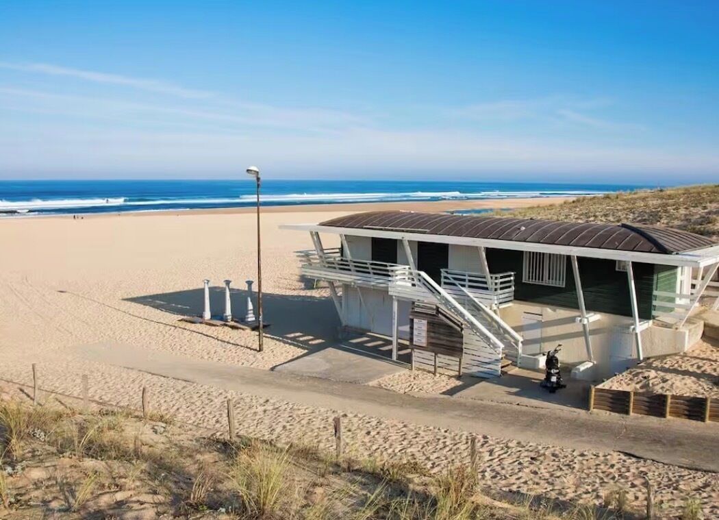 Un bâtiment situé au bord de la plage, avec un escalier menant à une zone abritée, une plage de sable en arrière-plan et un ciel dégagé.