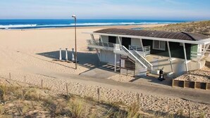 Un edificio frente a la playa con una escalera que lleva a un área cubierta, una playa arenosa al fondo y un cielo despejado.