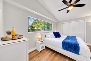 Une chambre à coucher comprenant un lit, une table de chevet, une commode, une fenêtre avec des stores, un ventilateur de plafond et une porte.