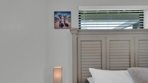 Hotellihuone, jossa on sänky, yöpöytä lampun kanssa, taulu seinällä ja ikkuna, jossa on sälekaihtimet.