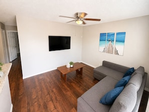 A living room with a flat-screen TV, a ceiling fan, a wooden coffee table, a grey sofa, and a beach-themed wall art. - Apartment - 3/4 Mile Walk to AT&T Stadium! (Arlington)