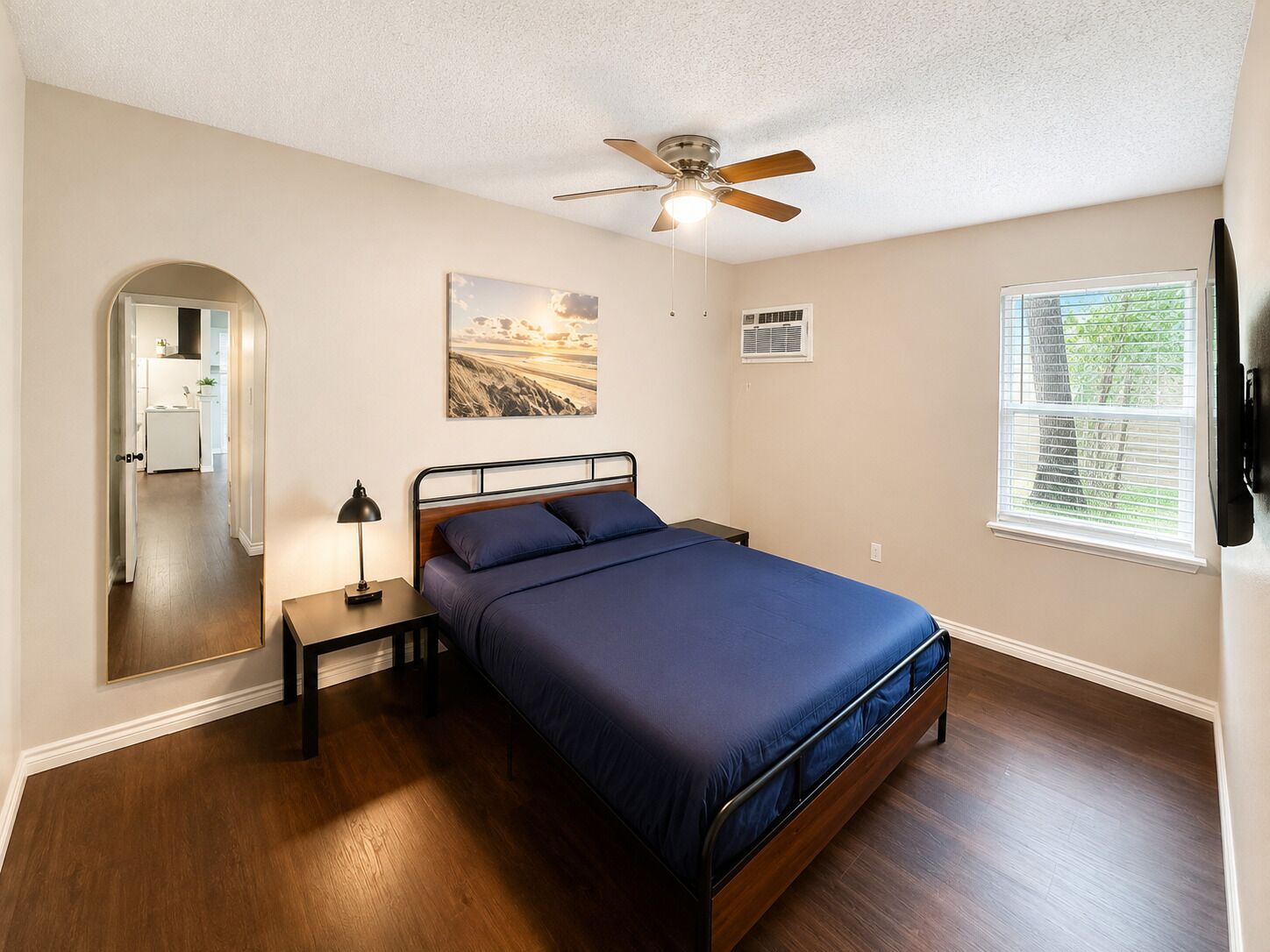 A bedroom with a bed, a nightstand, a ceiling fan, a window with blinds, and a door leading to another room.