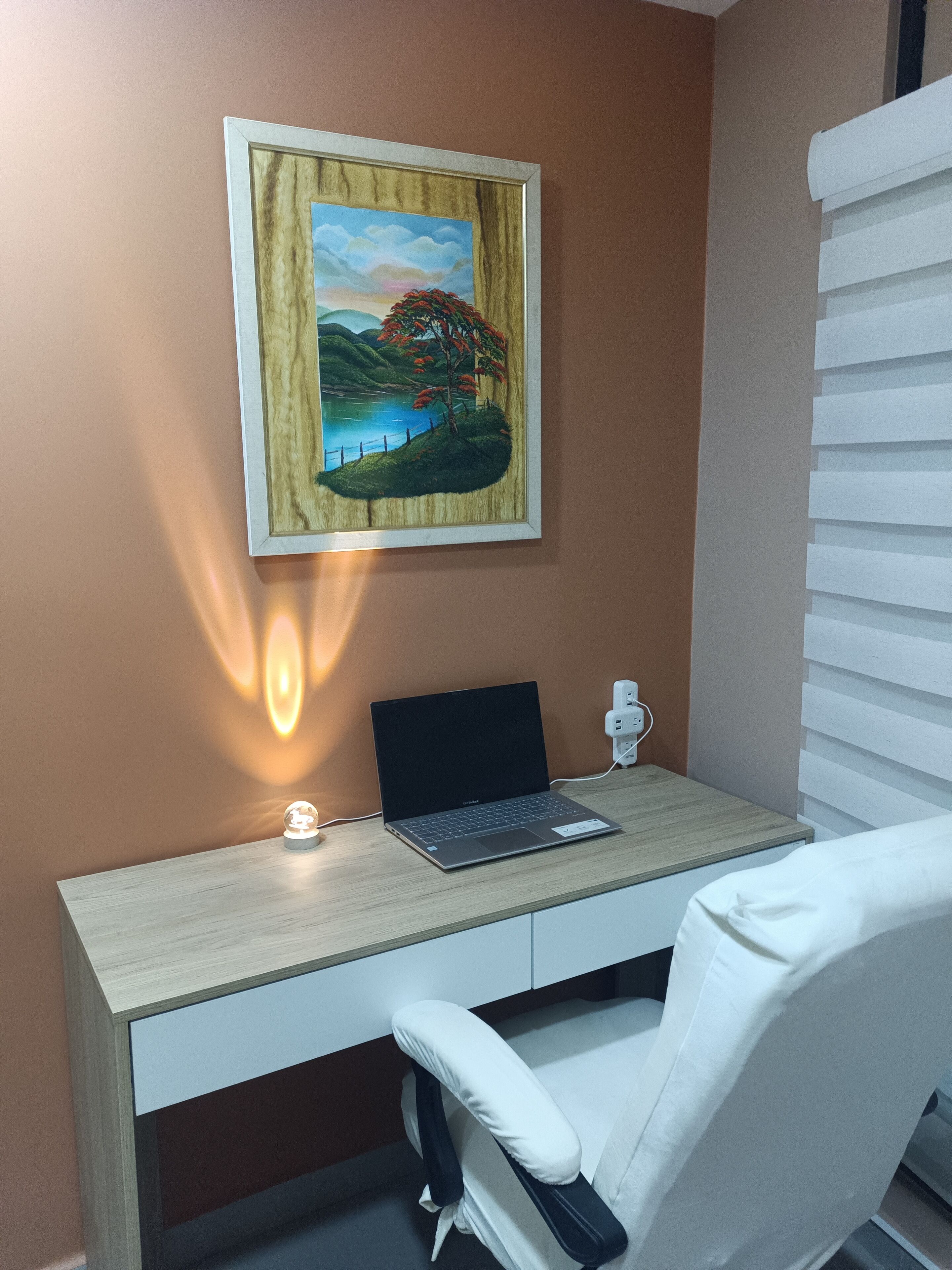 A room with a desk, a laptop, a chair, a wall-mounted lamp, and a painting.