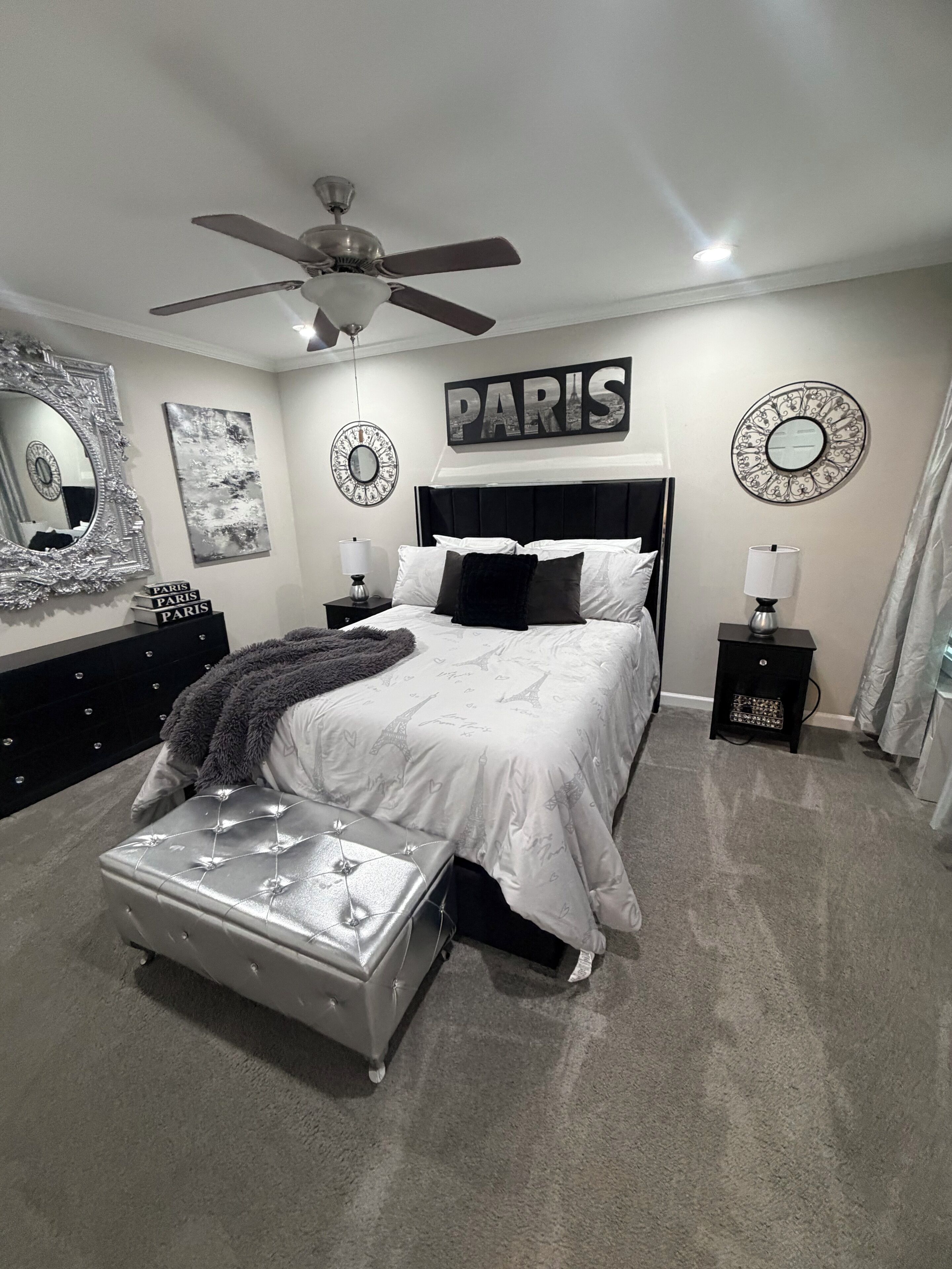 A bedroom with a bed, a ceiling fan, a nightstand with a lamp, a dresser, and decorative wall art.