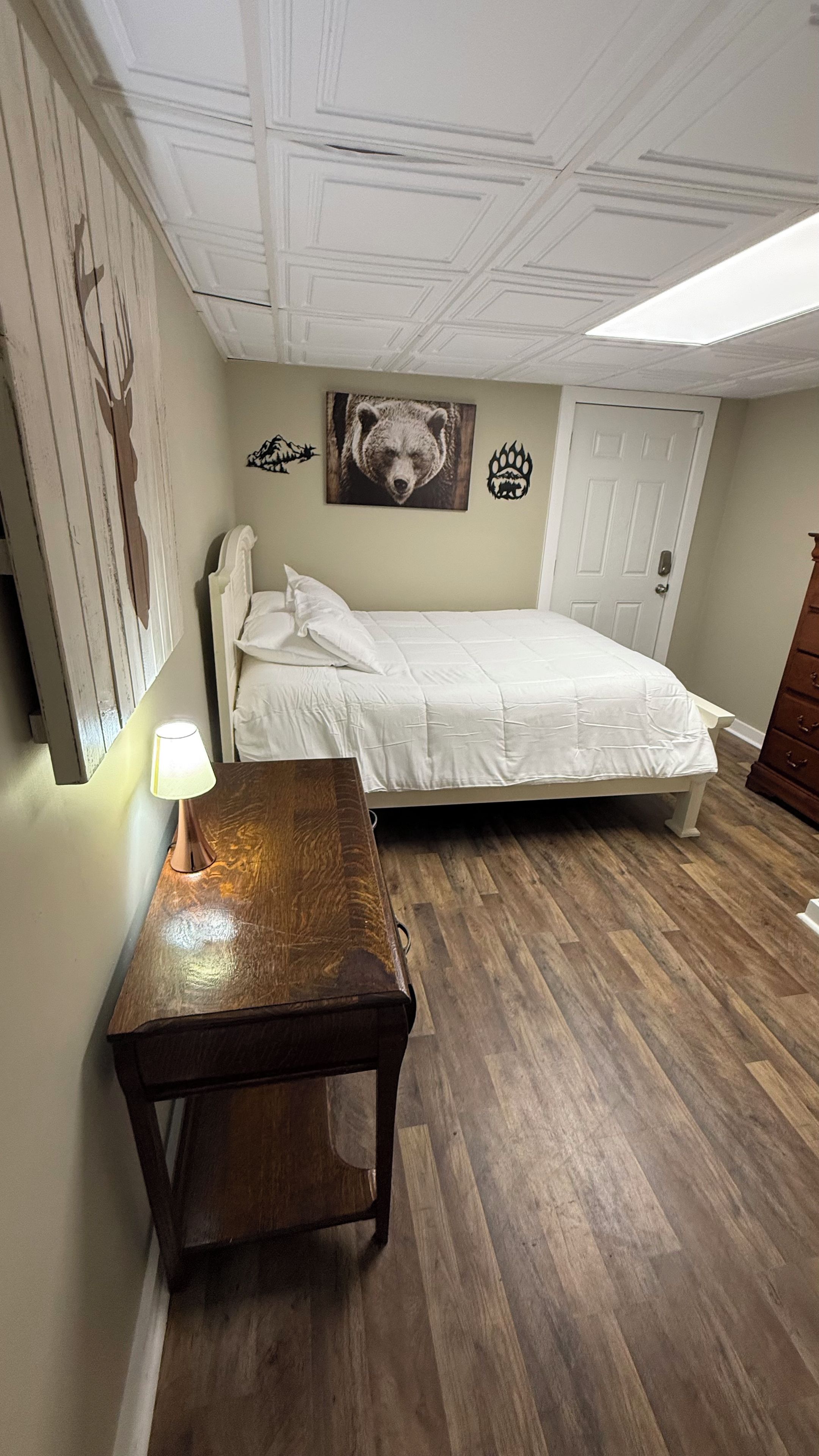 A bedroom with a bed, a bedside table with a lamp, a wooden dresser, and a wall with a bear painting and a paw print.