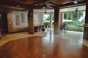 Una habitación amplia con piso de madera, un comedor con sillas y mesas, y ventanas grandes que ofrecen vistas a la vegetación exterior.