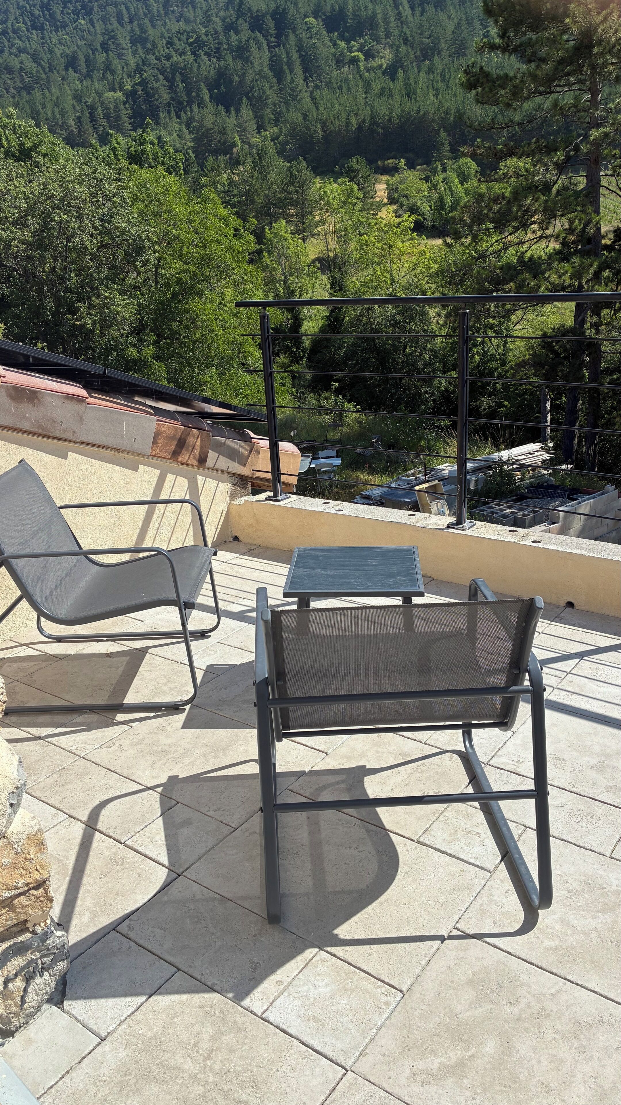 An outdoor patio with two metal chairs and a small table, surrounded by a low wall and a view of a forested hillside.