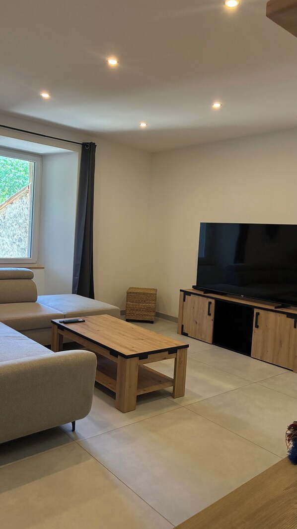 A modern living room with a sofa, a wooden coffee table, a television on a wooden stand, and a window with curtains.