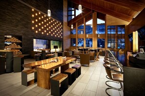 Eine moderne Bar mit Holztischen und Barhocker, großen Fenstern mit Blick ins Freie und einer Decke mit hängenden Lampen.