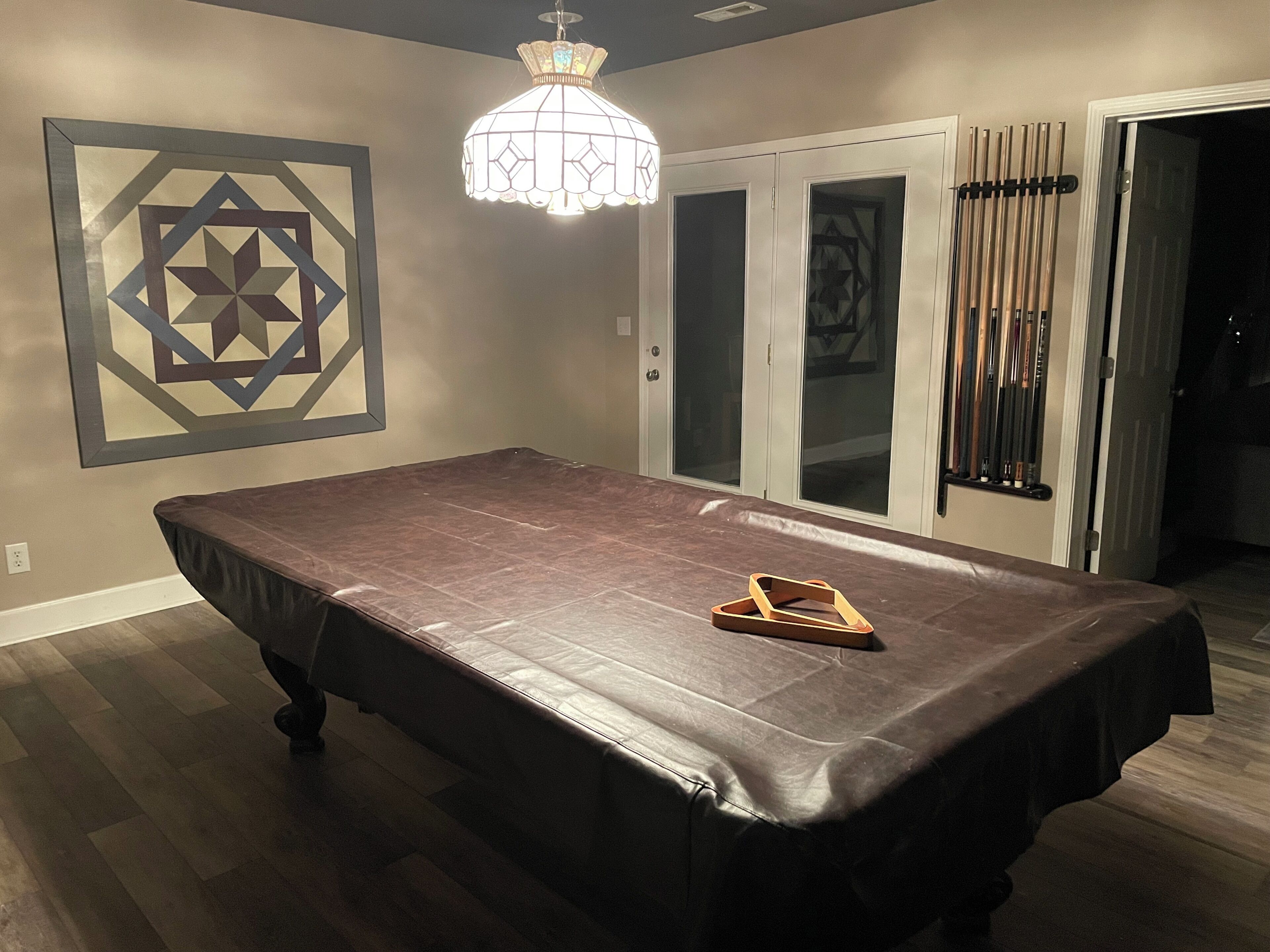 A pool table with a dark cloth, a geometric wall art piece, and a chandelier.