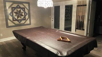 A pool table with a dark cloth, a geometric wall art piece, and a chandelier.