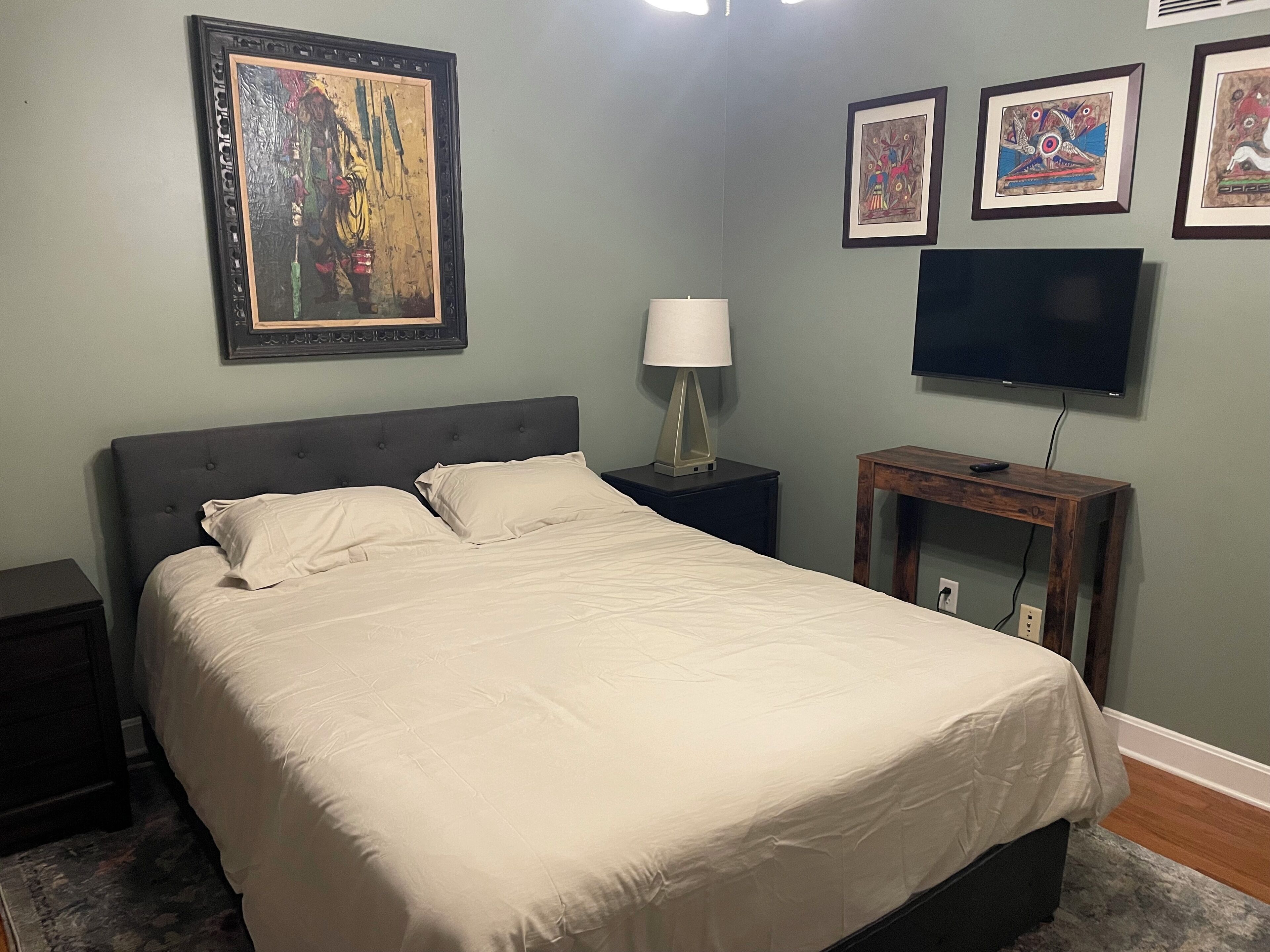 Un dormitorio con cama, mesita de noche, lámpara, televisor y cuadros en la pared.
