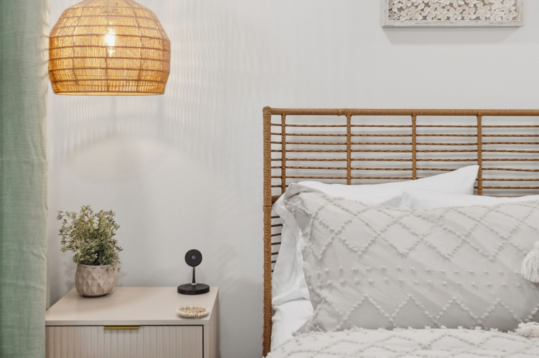 En seng med hvid dyne og et træhovedgærde, et natbord med en plante og en lampe, samt en væg med en dekorativ panel.