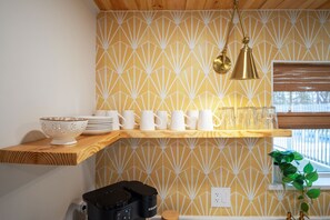 A kitchen with a wooden shelf holding white mugs and plates, a coffee maker, and a patterned wall.