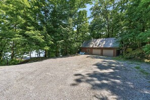A gravel driveway leading to a wooden cabin with a garage, surrounded by dense trees and a clear sky. - Seneca Lakefront: Sauna, Hot Tub & Game Room (Penn Yan)