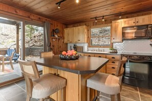 A modern kitchen with wooden cabinets, a central island with stools, a large oven, and a window overlooking a wooded area. - Seneca Lakefront: Sauna, Hot Tub & Game Room (Penn Yan)