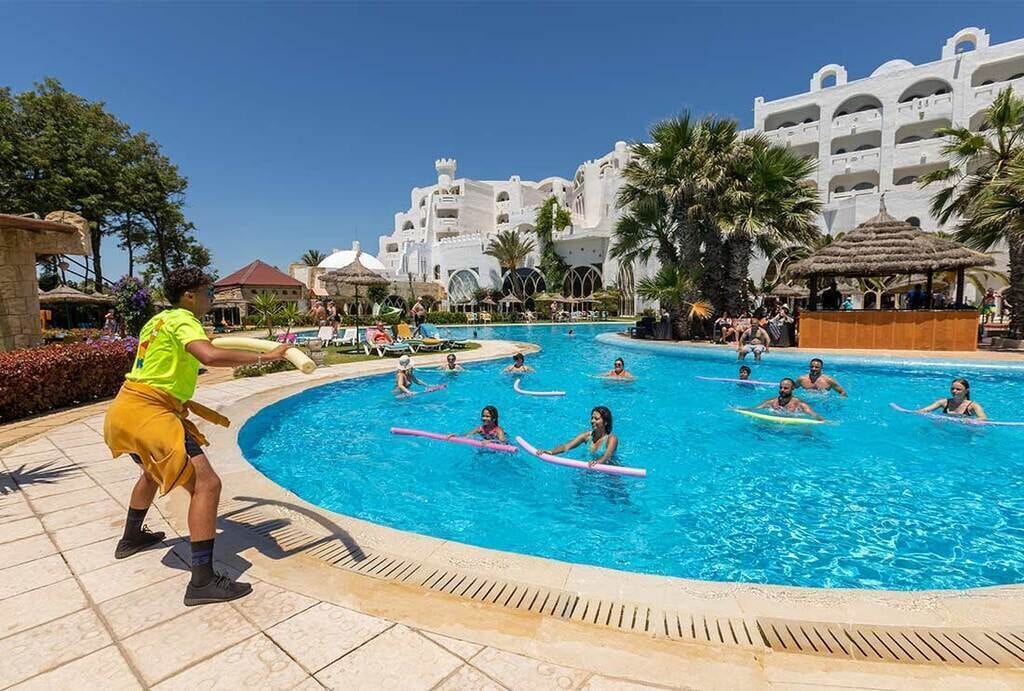A swimming pool area with multiple people, some on surfboards, and a person in a bright yellow outfit.