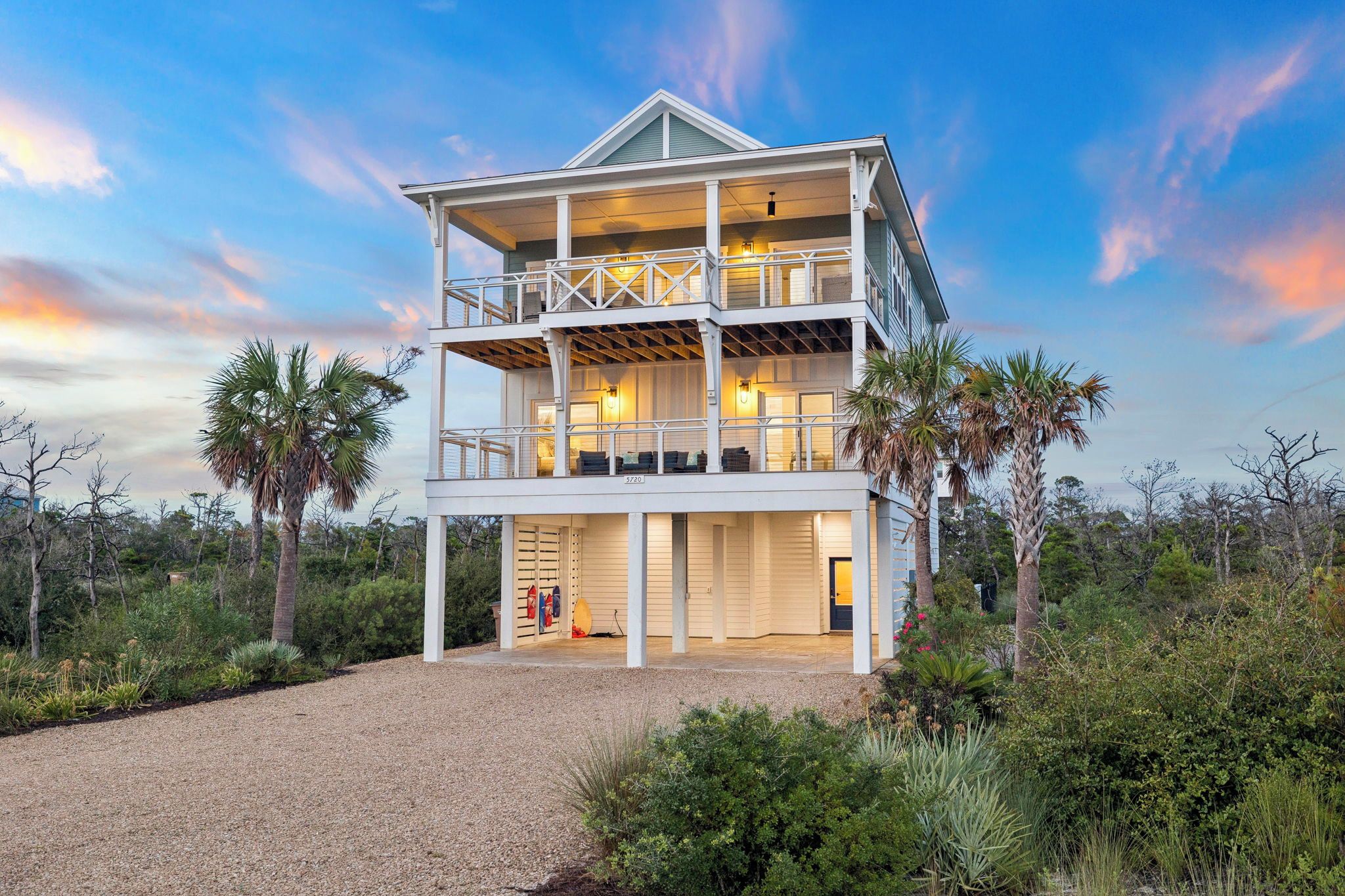 A multi-level beachfront property with a covered outdoor area, palm trees, and a gravel driveway.